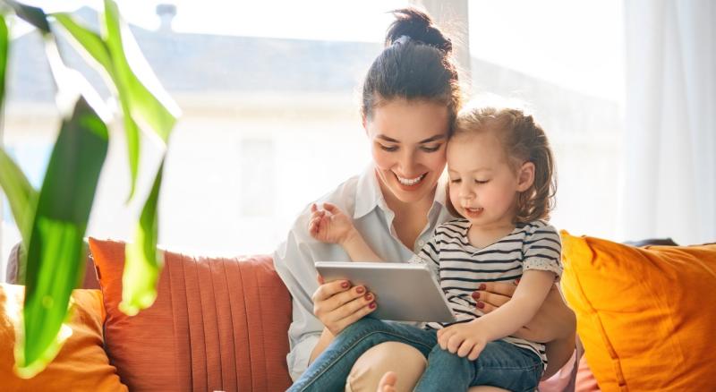 Woman and toddler, seated on couch, look at tablet.